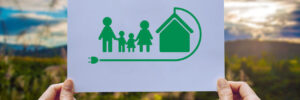Hands Holding Up Photo of Green Cartoon Family Next to Green House Eco-Friendly Concept
